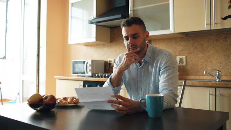 Person learning how to budget when living paycheck to paycheck, sitting at a kitchen table with bills and a notebook